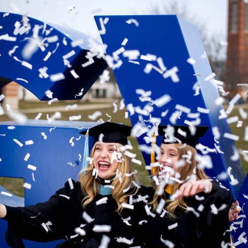Two graduates in caps and gowns laugh joyfully as white confetti falls around them in front of large blue letters.